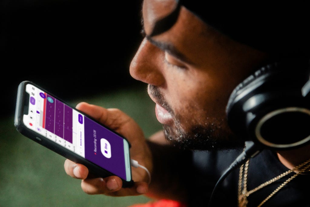 A man wearing headphones and speaking into a phone, the screen of the phone shows a voice recording app